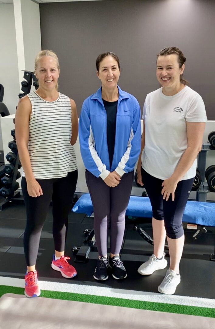 Three women standing in a gym with their arms crossed.
